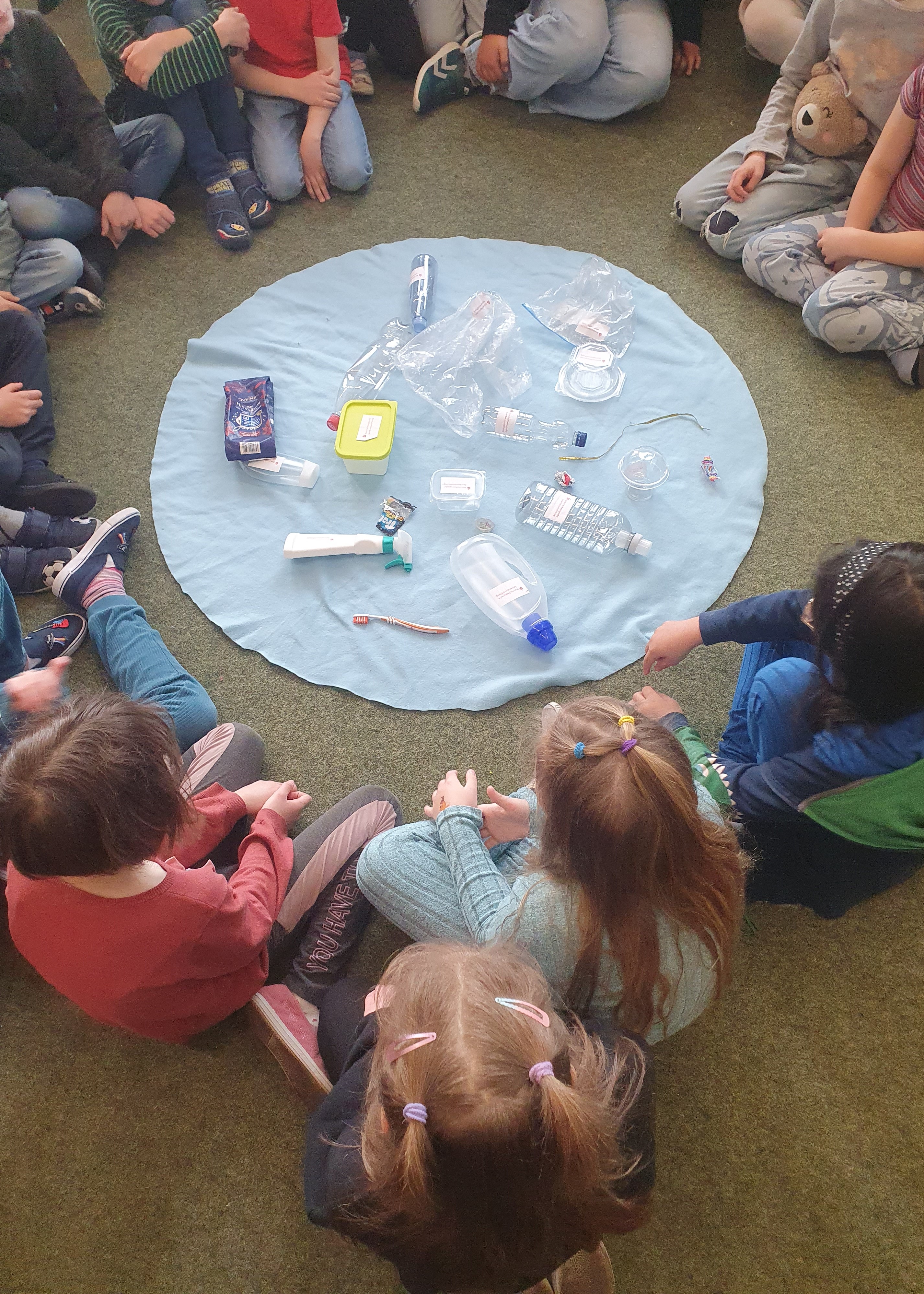 Kinder sitzen auf dem Boden um einen Teppich, auf dem Plastikmüll liegt.